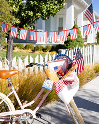 Pink bicycle with basket filled with gingham flag plates, floral scalloped plates, floral napkins, and American flags in front of white picket fence