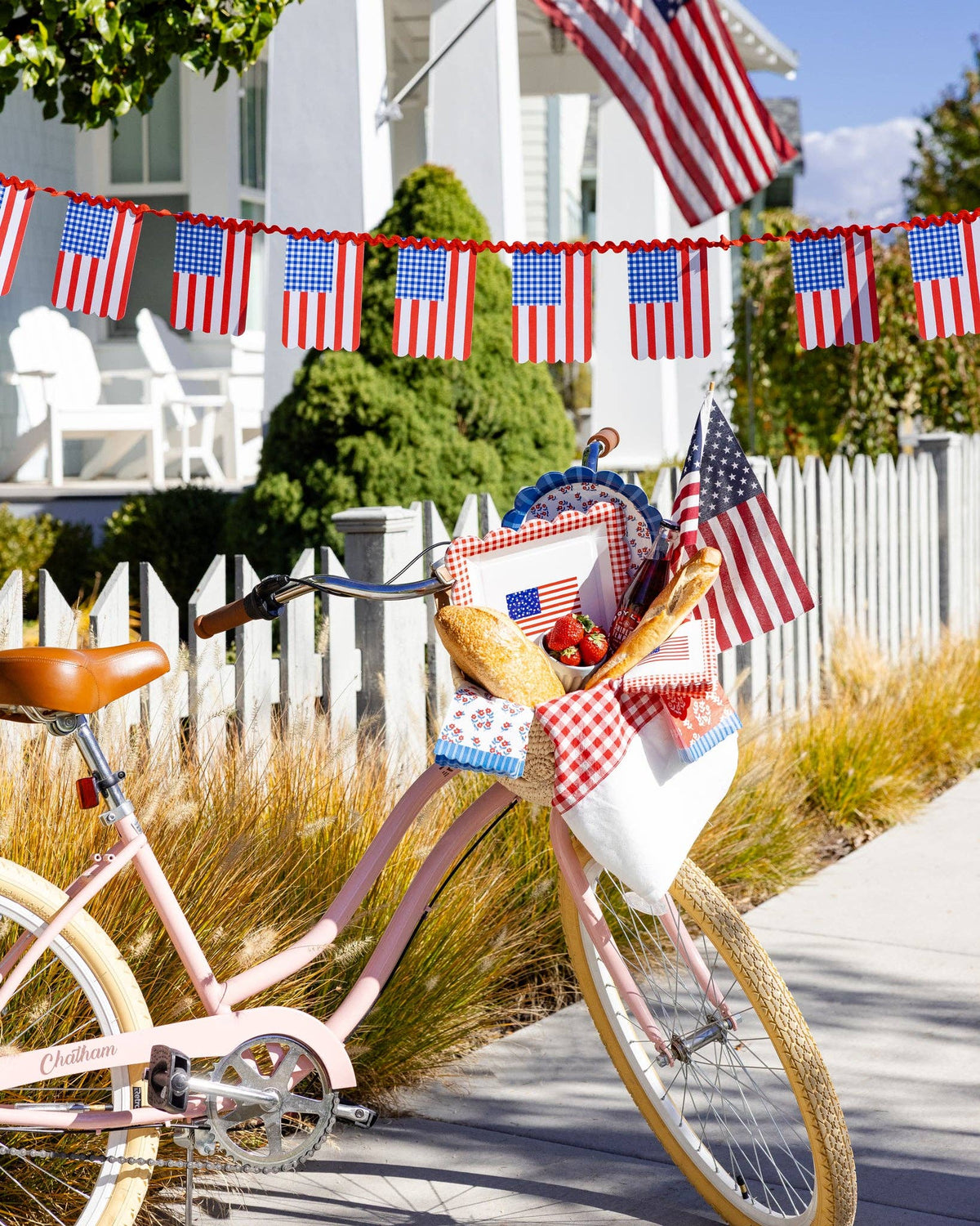 Americana gingham and stripe flag banner strung across a white picket fence with a pink bicycle and Fourth of July picnic basket in the foreground