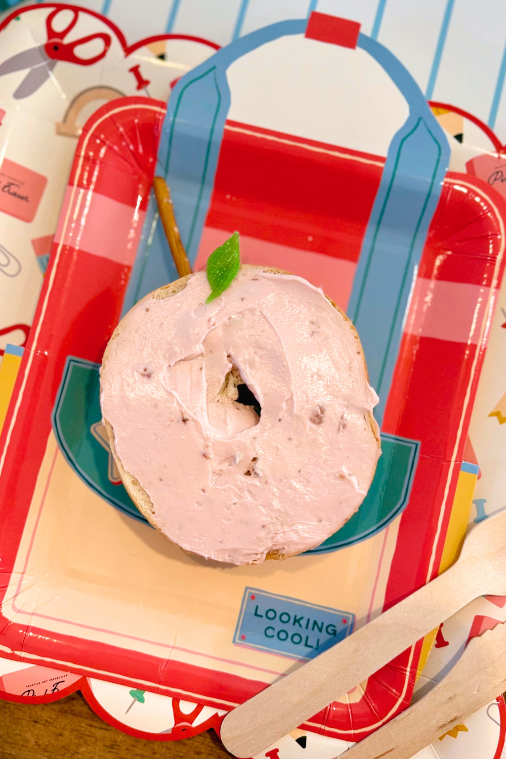 Apple-shaped bagel snack with strawberry cream cheese, green candy leaf, and pretzel stem served on a red backpack shaped plate layered over a back-to-school supplies scalloped paper plate with wooden utensils.