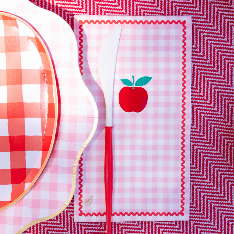 Detail close-up of pink gingham apple paper guest towel beside a red apple shaped paper plate and white plastic cutlery with red handle on a red chevron tablecloth, showing the rickrack border and central apple illustration