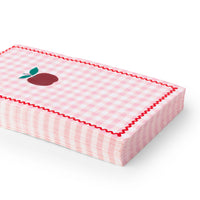 Close-up side view of stack of pink gingham apple paper guest towels showing folded napkin stack with red rickrack border, red apple illustration with green leaves, and pink gingham pattern