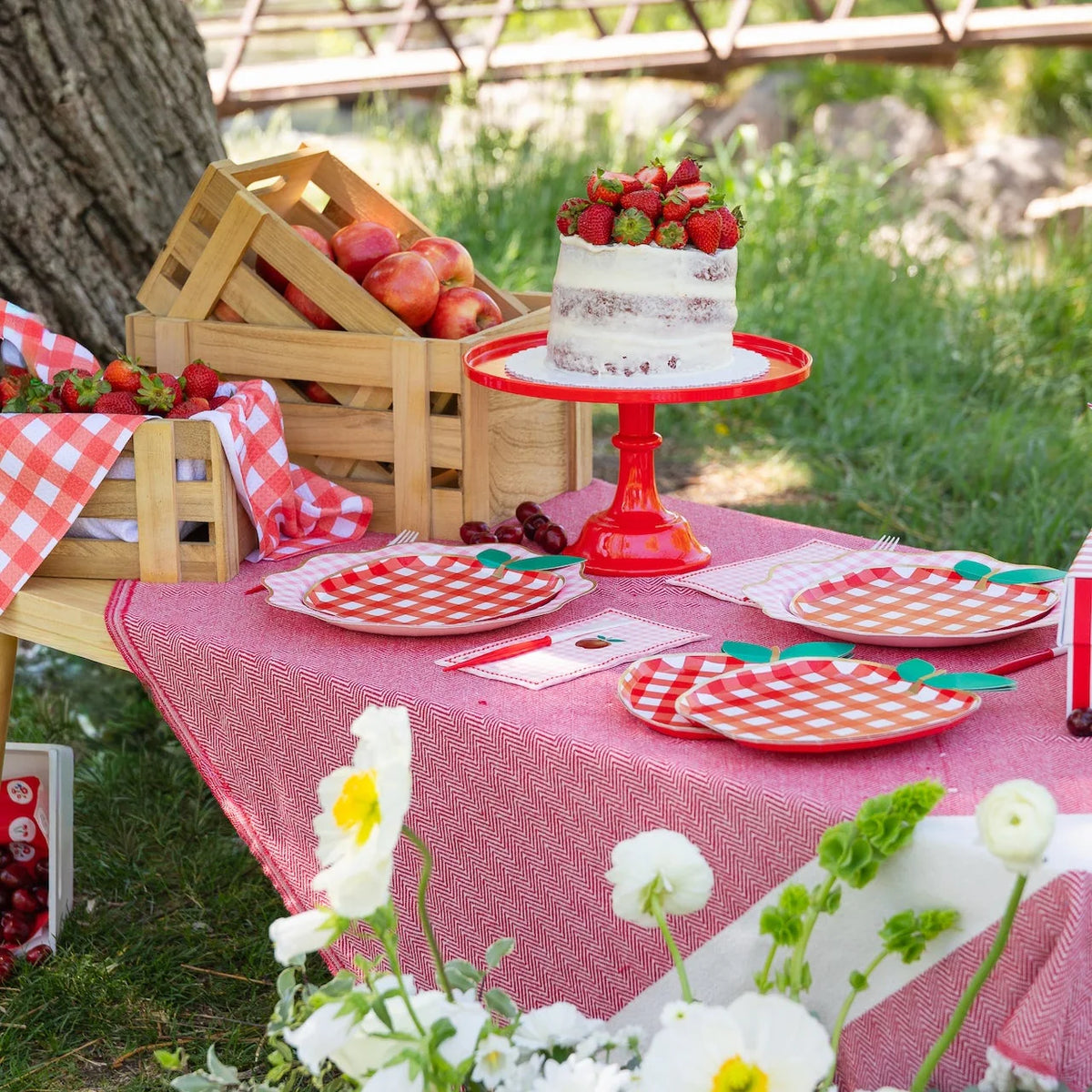 Outdoor summer picnic tablescape under a tree with red apple shaped paper plates layered on top of red and white gingham paper plates, pink gingham apple paper guest towels, red plastic cutlery, strawberry-topped cake on red cake stand, and wooden crates of apples and strawberries