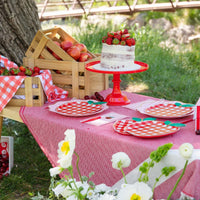 Outdoor summer picnic tablescape under a tree with red apple shaped paper plates layered on top of red and white gingham paper plates, pink gingham apple paper guest towels, red plastic cutlery, strawberry-topped cake on red cake stand, and wooden crates of apples and strawberries