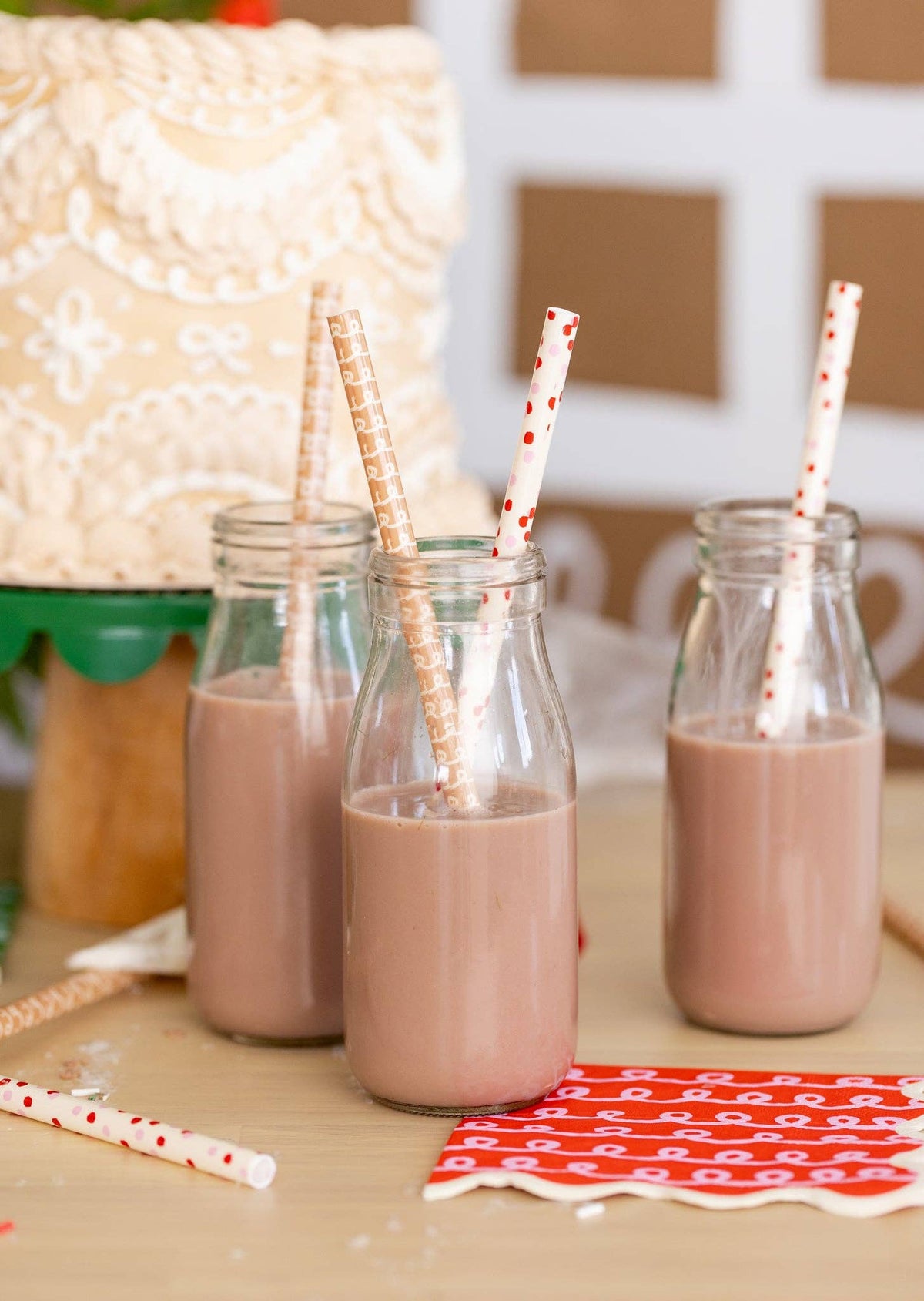 Three glass bottles of chocolate milk with Gingerbread Straws beside a frosted holiday cake and cookies.