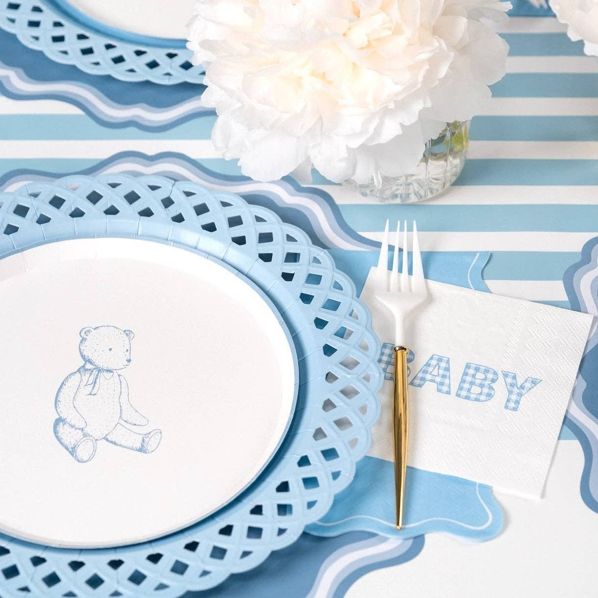 Styled baby shower place setting close-up showing the teddy-bear plate on a blue scalloped charger with a "BABY" napkin and blue-striped runner.