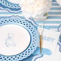 Styled baby shower place setting close-up showing the teddy-bear plate on a blue scalloped charger with a "BABY" napkin and blue-striped runner.