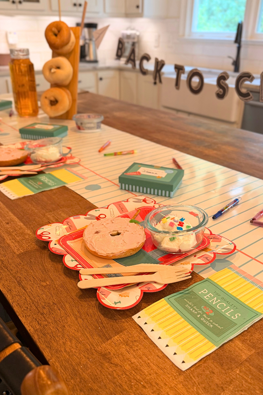 Angled view of a styled back-to-school breakfast table with a bagel apple snack on a red backpack shaped plate, layered over a back-to-school supplies scalloped paper plate, with pencil box napkins, lunchbox treat boxes, and colorful school-themed details.