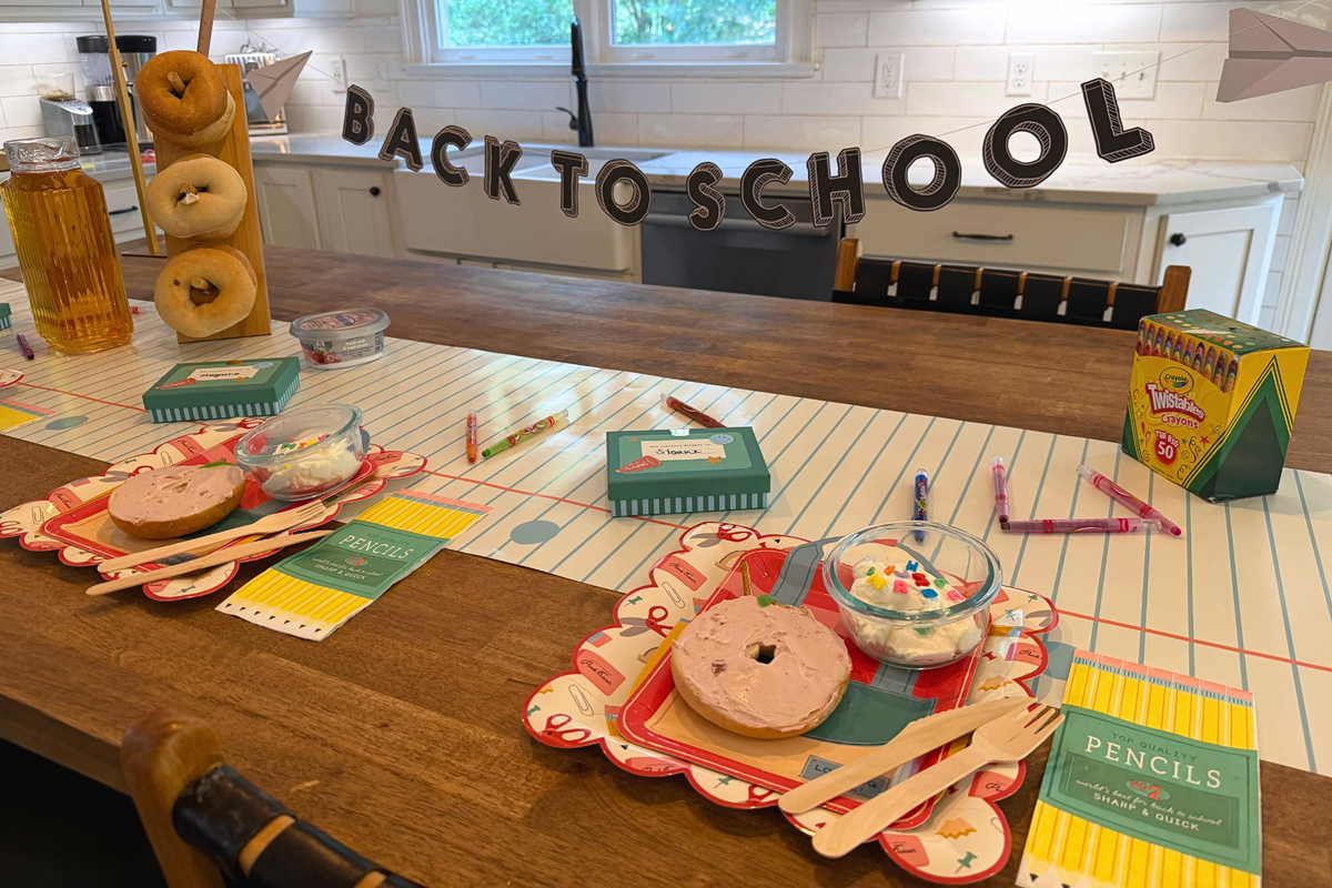 Wide back-to-school breakfast tablescape with two place settings featuring red backpack shaped plates layered over back-to-school supplies scalloped paper plates, bagel apple snacks, pencil box napkins, lunchbox treat boxes, and colorful school supplies.