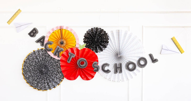 Back to School chalkboard-letter banner strung with paper airplanes and rulers, hung across paper fans, an apple, and school-themed party decor on a white wall.