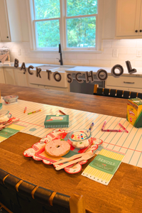 Back-to-school breakfast table featuring a red backpack shaped plate layered over a back-to-school supplies scalloped paper plate, bagel apple snack, pencil box napkin, lunchbox treat boxes, colorful school supplies, and a back-to-school banner on a notebook paper table runner.