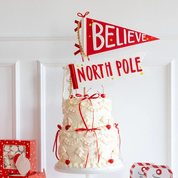 Layered cake topped with Believe and North Pole felt pennant flags, styled with red and white Christmas party supplies.