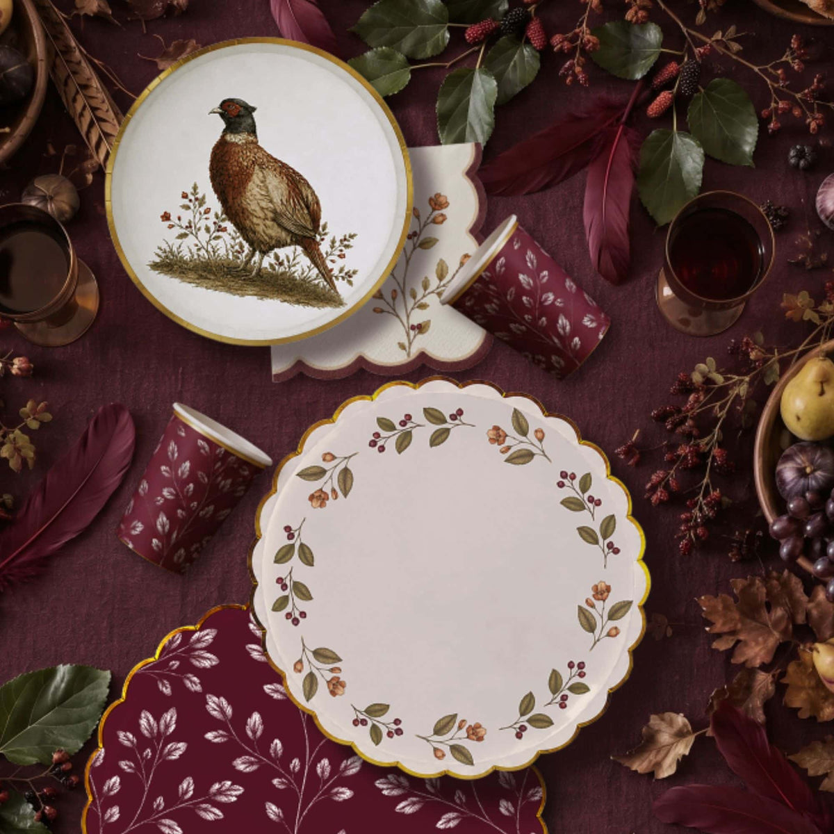 Fall flat lay with a pheasant plate, berry wreath plate, burgundy leaf cups, and burgundy napkins on plum linen