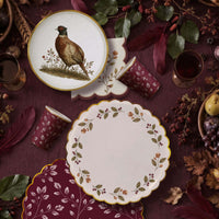 Fall flat lay with a pheasant plate, berry wreath plate, burgundy leaf cups, and burgundy napkins on plum linen