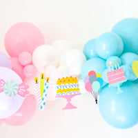 Pastel balloon arch with “It’s a Birthday” garland hanging across—die-cut cakes, candles, candy and balloons create a candy-land party backdrop.