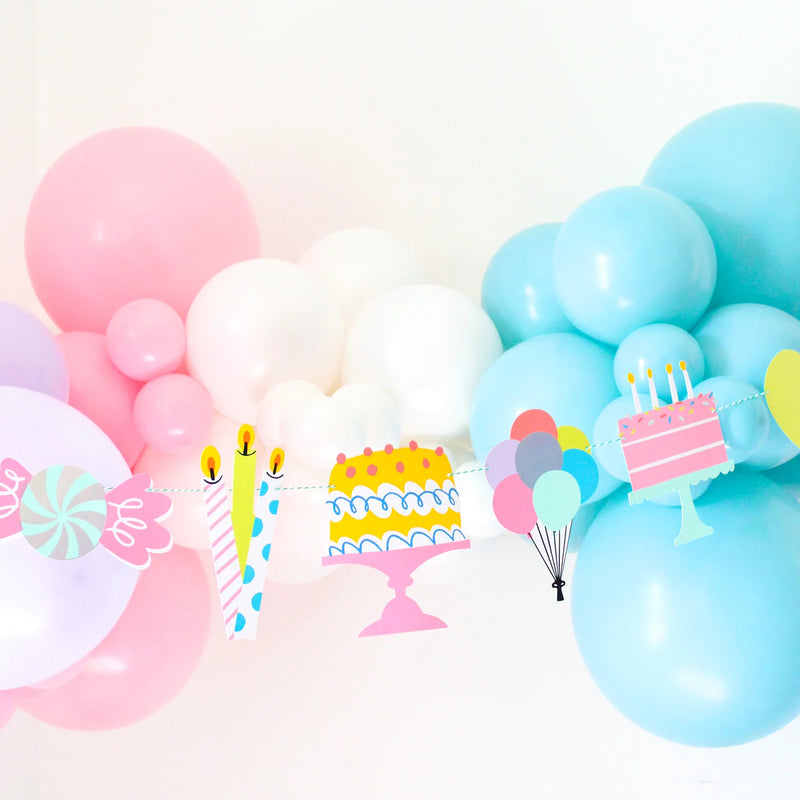 Pastel balloon arch with “It’s a Birthday” garland hanging across—die-cut cakes, candles, candy and balloons create a candy-land party backdrop.