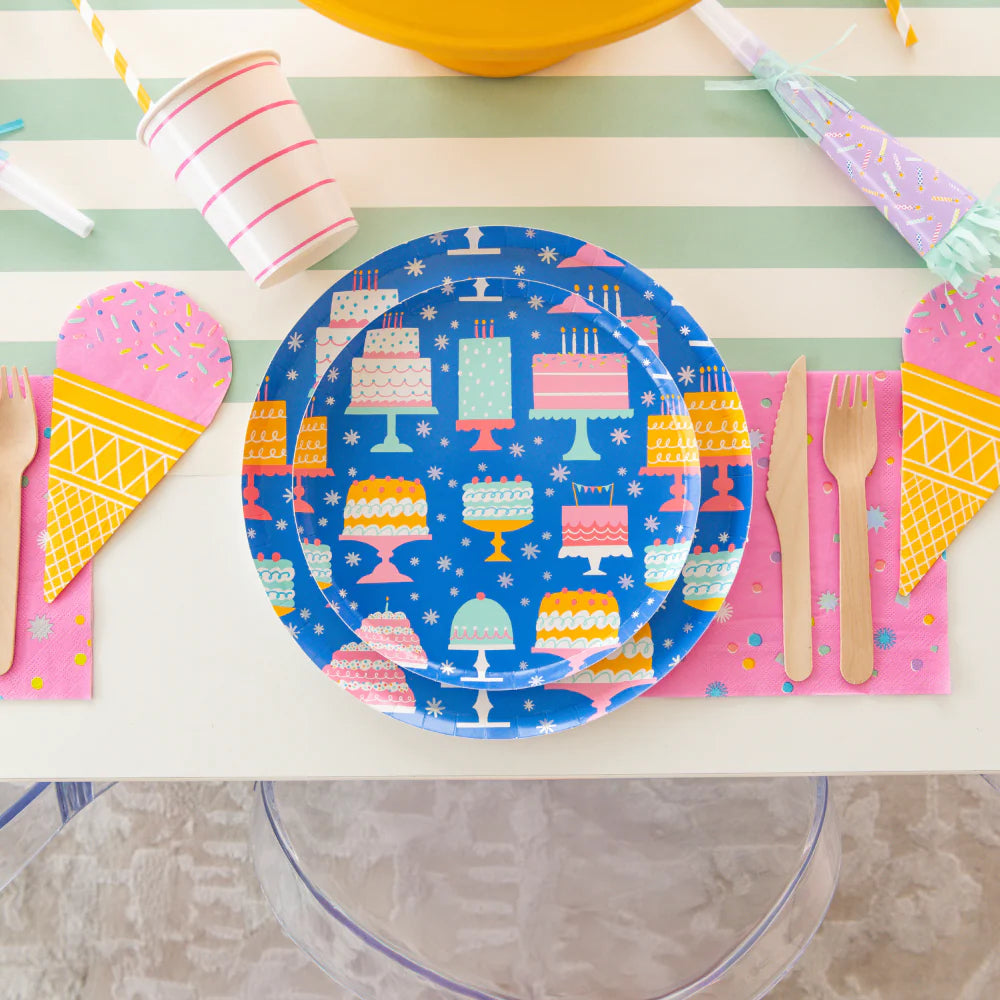 Close-up of a place setting: blue birthday-cake plate, pink confetti napkin, wooden cutlery and ice-cream cone décor on striped runner.