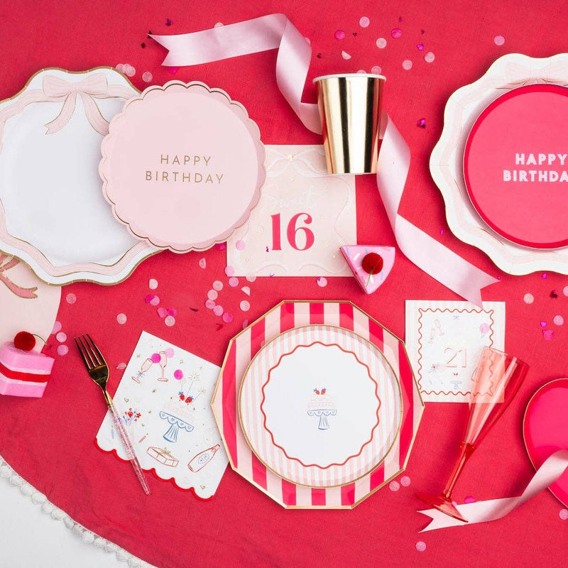 Overhead lifestyle flat lay of pink girls birthday party table with striped birthday cake plates, bow plates, illustrated celebration napkins, gold cup and confetti on red tablecloth