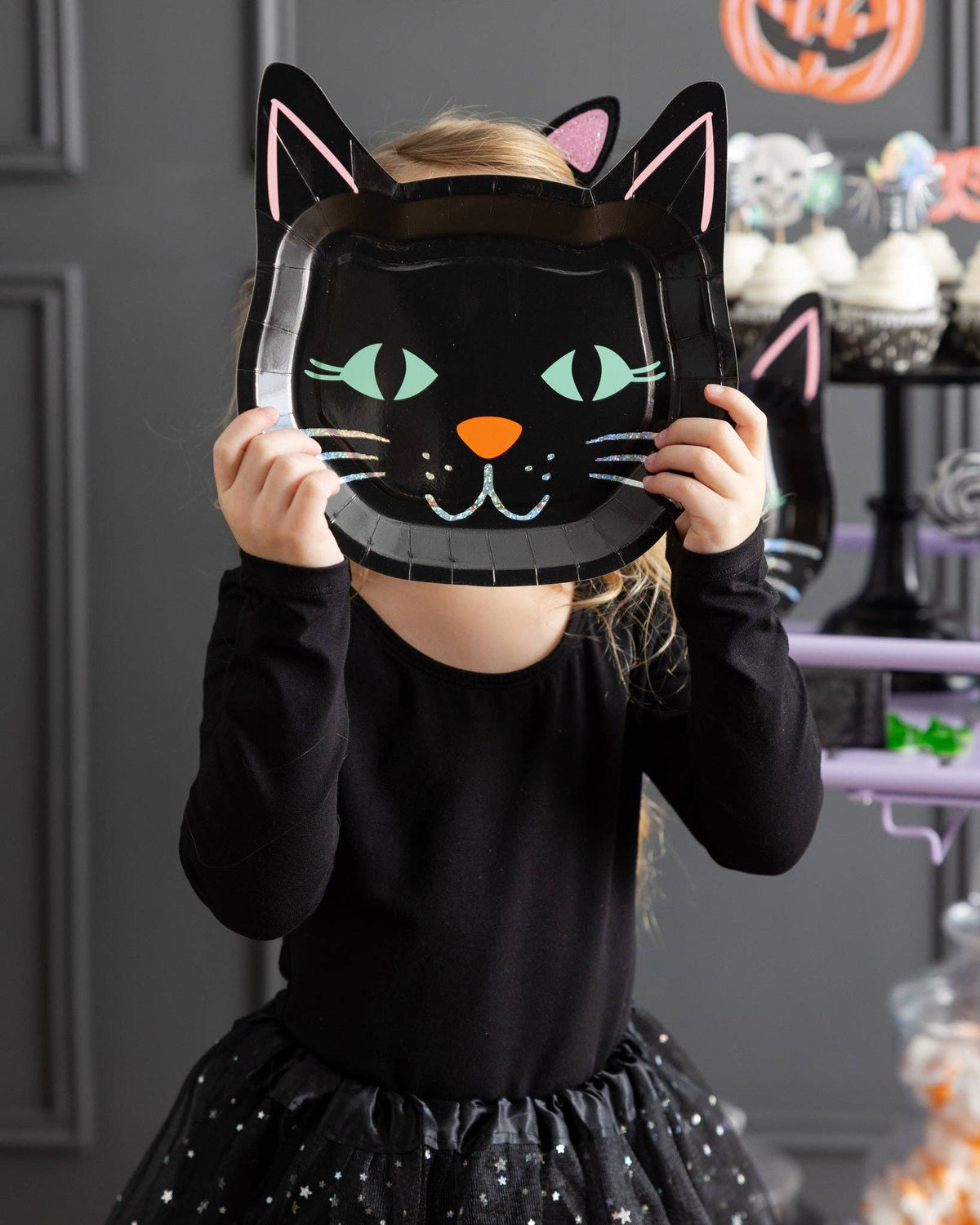 Close-up of the girl holding the black cat plate as a face mask, showing the colorful cat features and holographic glow.