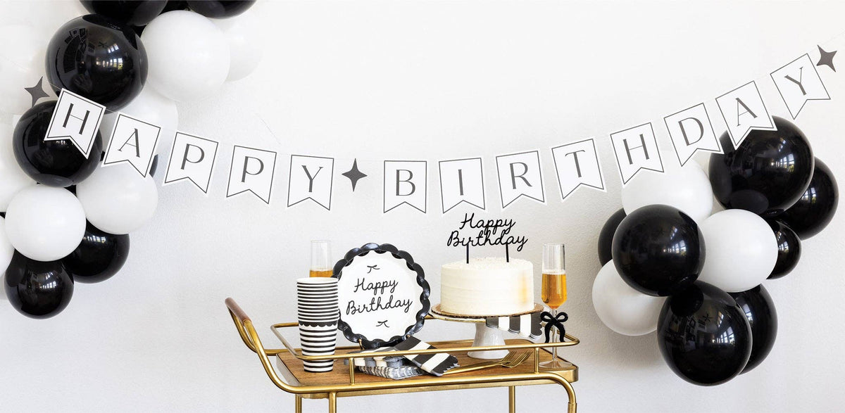 Black and cream Happy Birthday banner hung above gold bar cart with cake, black and white balloons, and coordinating party supplies