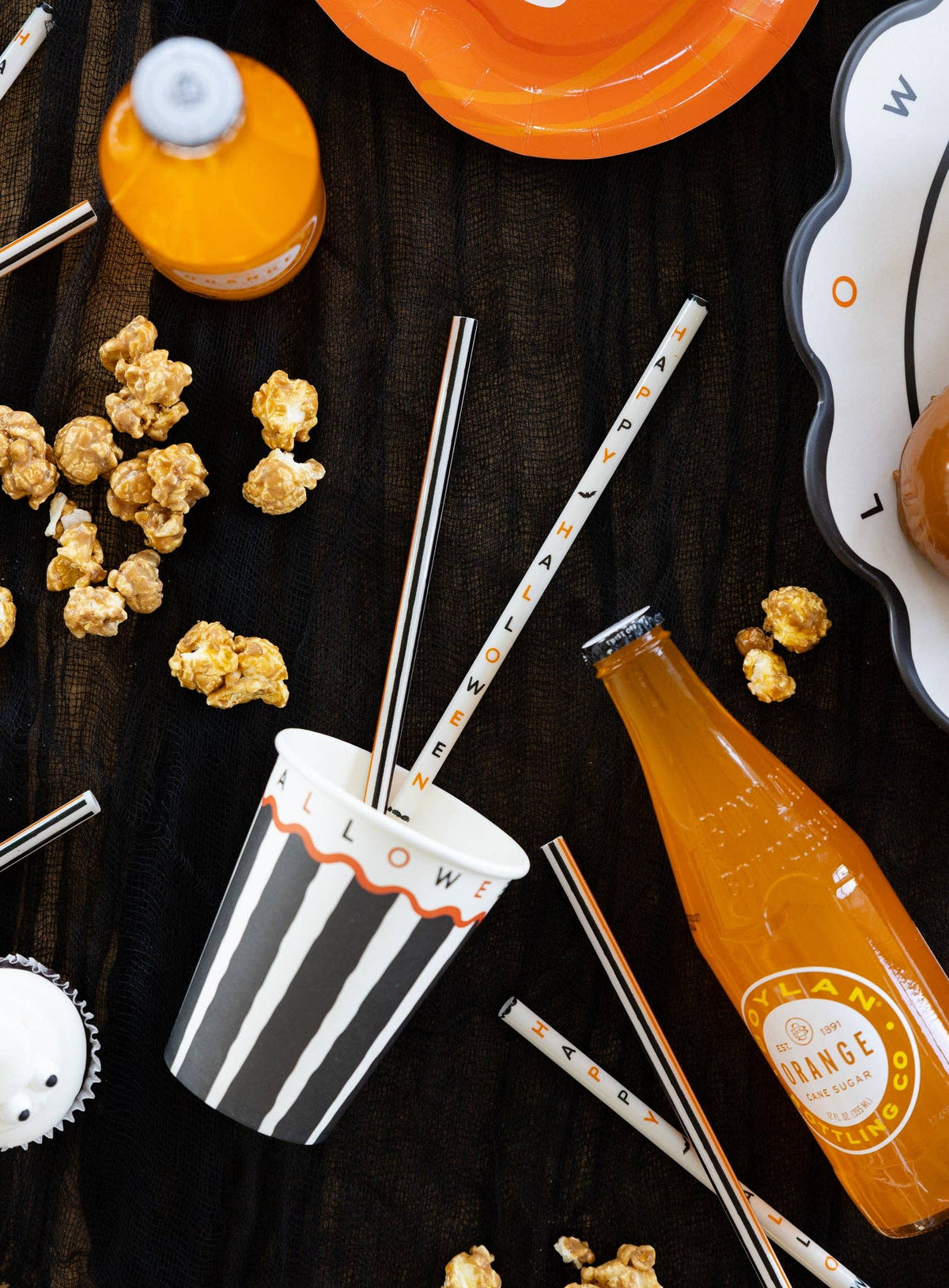 Halloween-themed drinks with striped cups, orange bottles, popcorn, and the Happy Halloween reusable straws on a wooden surface.
