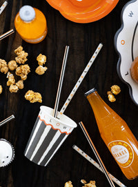 Halloween-themed drinks with striped cups, orange bottles, popcorn, and the Happy Halloween reusable straws on a wooden surface.
