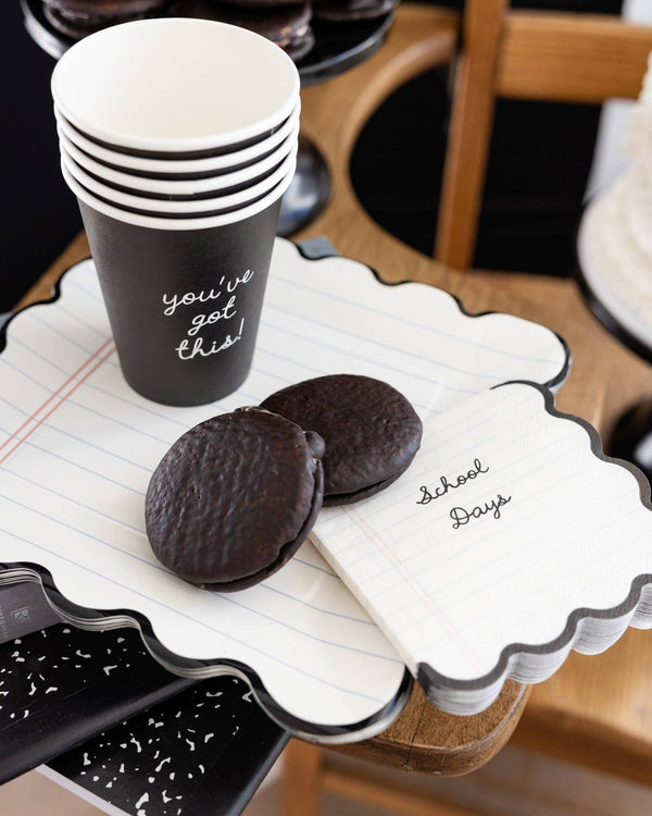 Close-up of single black "you've got this!" paper party cup stacked with additional cups on a notebook paper scalloped plate, with chocolate cookies alongside