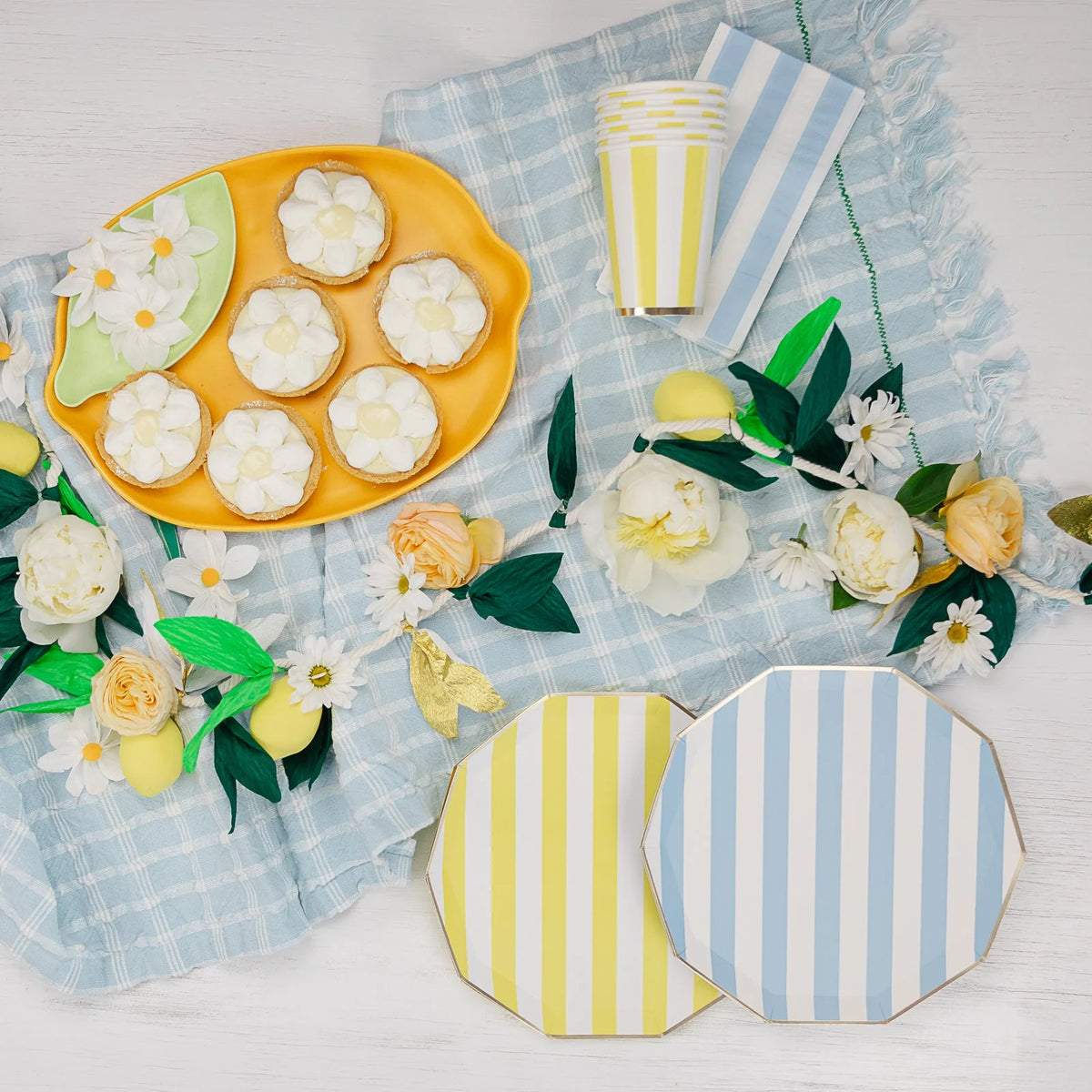 Blue cabana stripe paper dinner plates and yellow cabana stripe plates styled on a blue plaid throw with a citrus party tablescape, paper flower garland, and lemon tart desserts