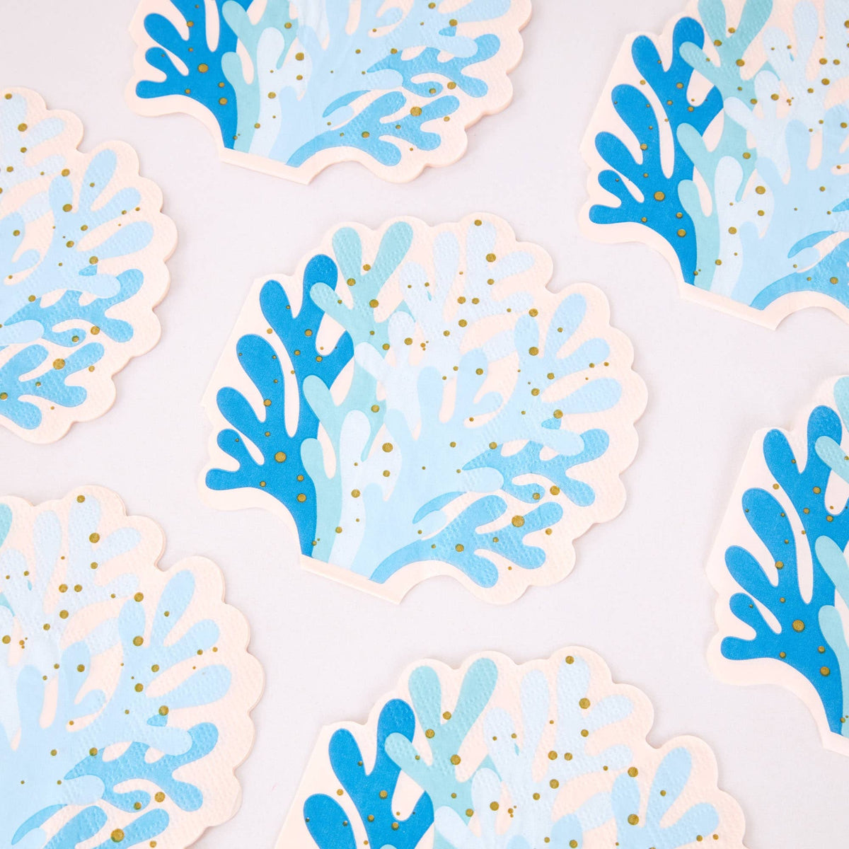 Blue coral shaped paper cocktail napkins scattered, showing the die-cut reef silhouette and gold foil speckles.