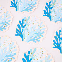 Blue coral shaped paper cocktail napkins scattered, showing the die-cut reef silhouette and gold foil speckles.