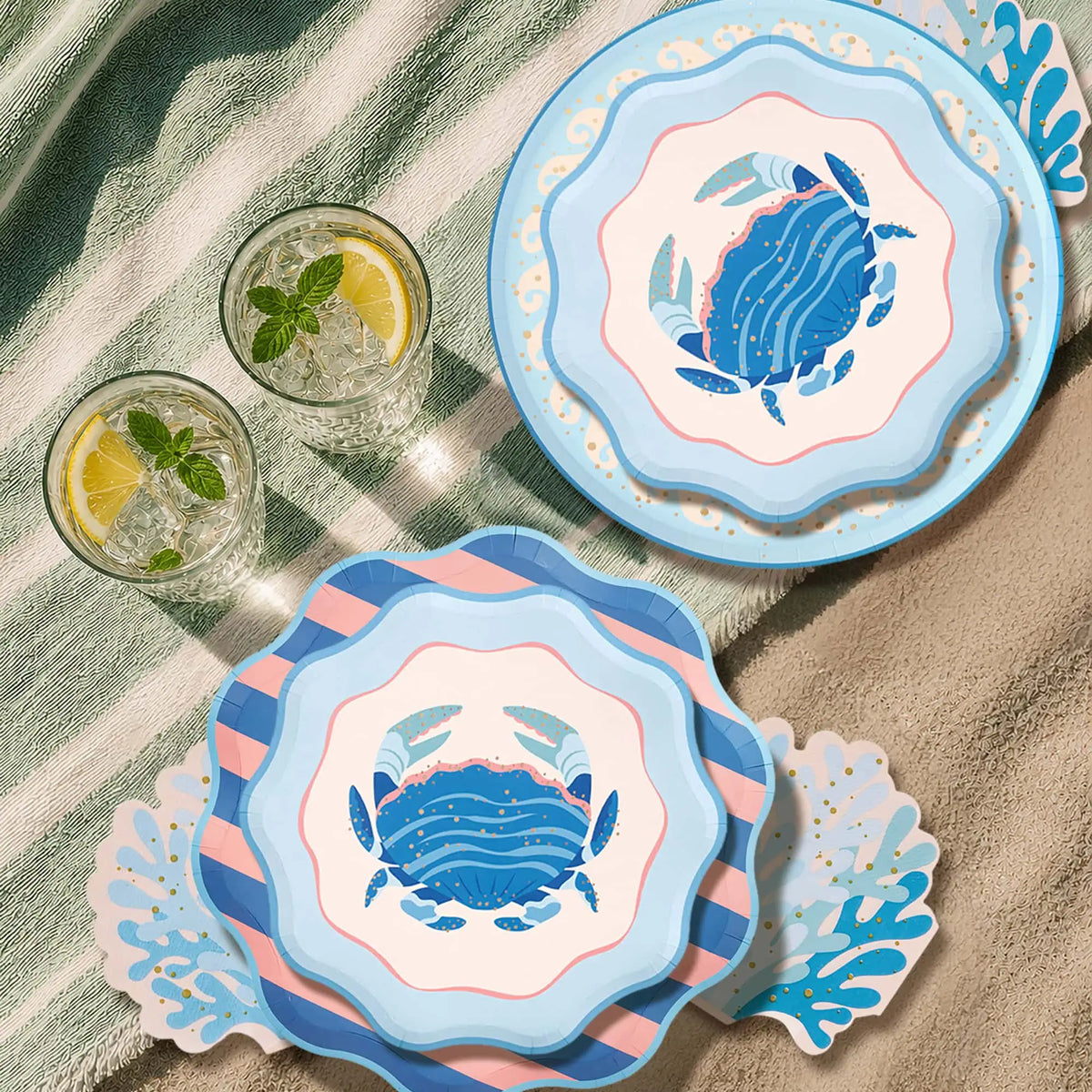 Outdoor lifestyle: the round blue crab dinner plate and the scalloped blue crab salad plate stacked over a striped plate on a green-and-white striped towel and sand, two iced drinks with lemon and mint, and blue coral-shaped napkins. Overhead styled shot.