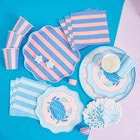 Full party flatlay on blue and pink ground: pink/blue striped cups, pink/blue and blue/white striped scalloped plates, striped napkins, blue coral-shaped napkins, the round crab dinner plate, the scalloped crab salad plate, and macarons. Overhead collection shot.