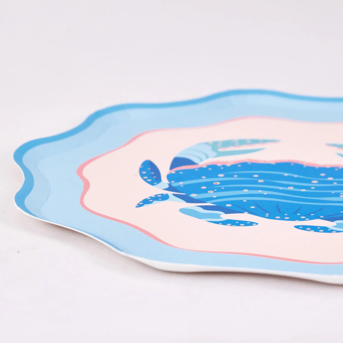Angled edge detail of a single scalloped blue crab plate on white, highlighting the layered light-blue rim, pink ring, blush field, and gold-flecked crab. Close-up product shot.