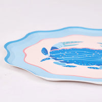 Angled edge detail of a single scalloped blue crab plate on white, highlighting the layered light-blue rim, pink ring, blush field, and gold-flecked crab. Close-up product shot.