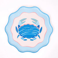 A single scalloped blue crab plate, straight-on overhead on a white background: cobalt crab with light-blue claws and gold foil dots, blush center, pink ring, light-blue scalloped rim. Clean hero product shot.