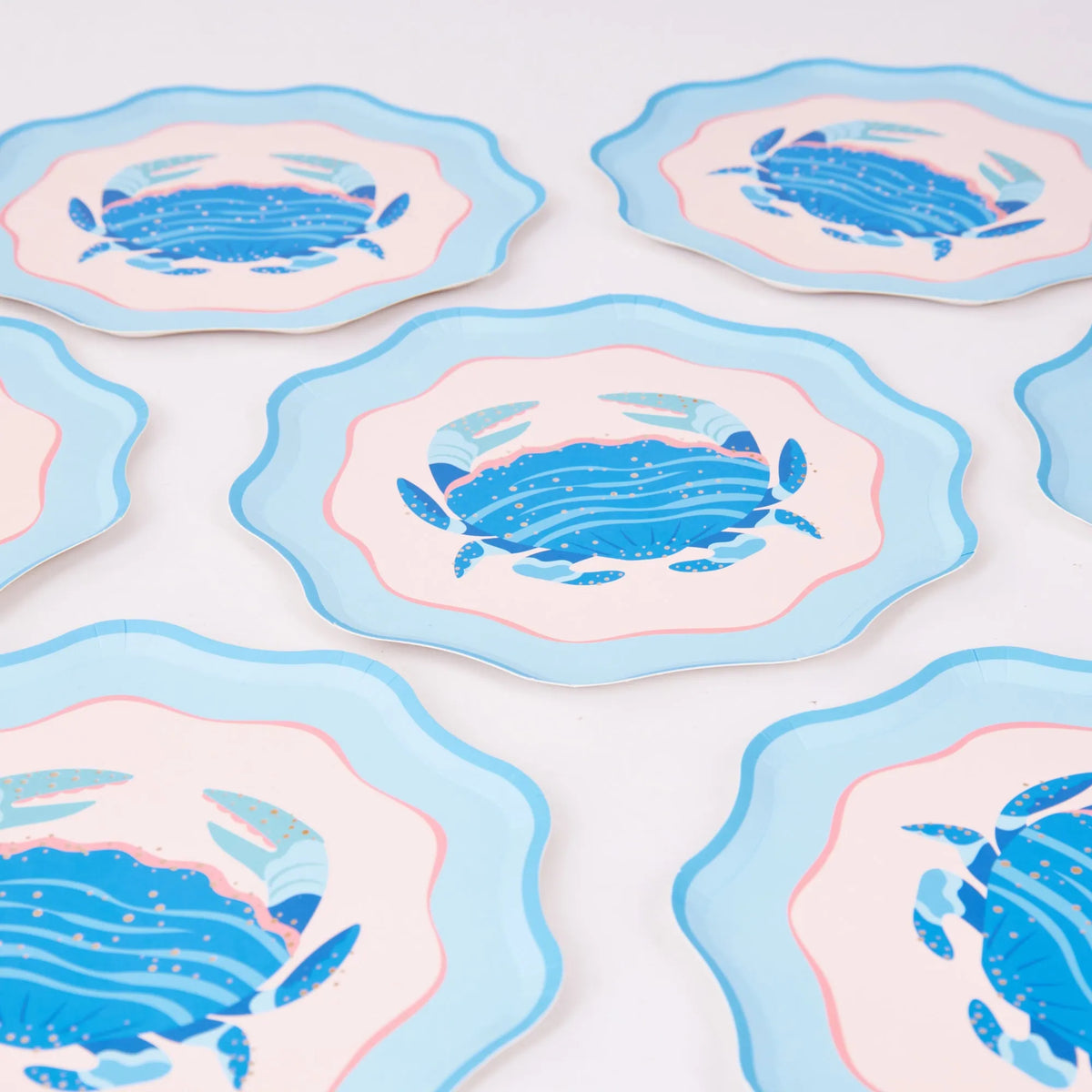 A scattered group of the scalloped blue crab salad plates on a white background, each showing the cobalt crab, blush center, pink ring, and light-blue wavy rim from slightly different angles. Overhead product shot.