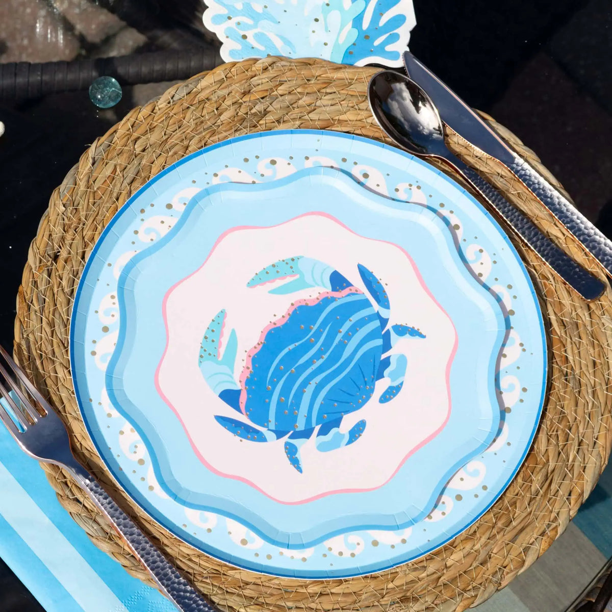 Overhead place setting on a round woven seagrass charger: the blue crab scalloped salad plate layered over a light-blue round crab plate with a white swirl-wave gold-dot border, rose-gold flatware alongside, a blue striped napkin, and a blue wave-shaped die-cut at the top edge. Sunlit angled lifestyle shot.