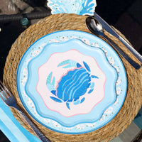 Overhead place setting on a round woven seagrass charger: the blue crab scalloped salad plate layered over a light-blue round crab plate with a white swirl-wave gold-dot border, rose-gold flatware alongside, a blue striped napkin, and a blue wave-shaped die-cut at the top edge. Sunlit angled lifestyle shot.