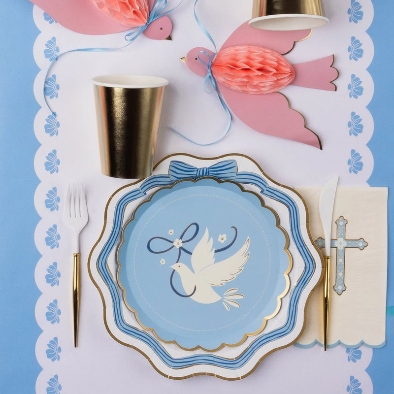 Close-up of a blue dove scalloped paper plate featuring a white dove with ribbon and flowers, layered over a blue striped bow plate on a scalloped floral placemat with a cross napkin, gold flatware, gold paper cup, and pink honeycomb dove decoration.