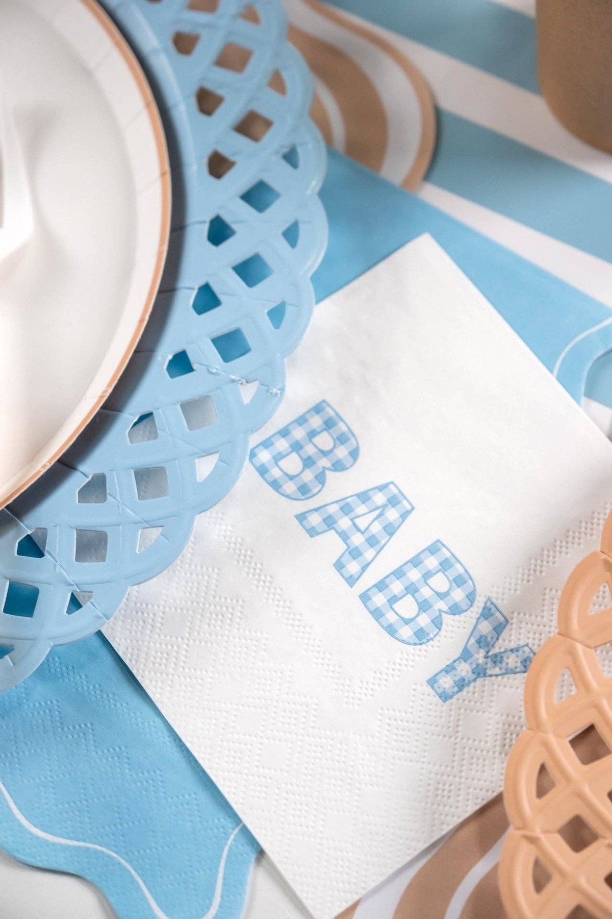Blue gingham BABY napkin on a blue and tan striped table with a lattice charger and kraft cup