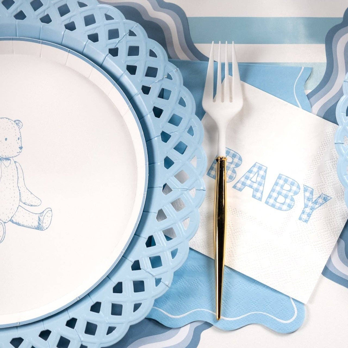 Blue gingham BABY napkin beside a teddy bear plate on a blue lattice charger with a gold handled fork