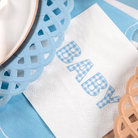 Close view of the BABY lettering in blue gingham on a white napkin beside blue and tan lattice chargers