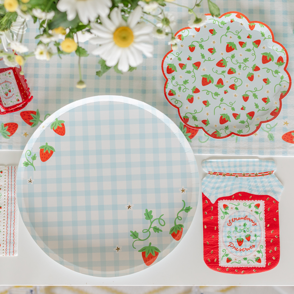 Berry first birthday place setting featuring the blue gingham strawberry paper plate, white and red striped paper cup, strawberry stripe cocktail napkin, wooden cutlery, fresh strawberries in a green pint basket, and a wildflower bouquet with daisies and chamomile
