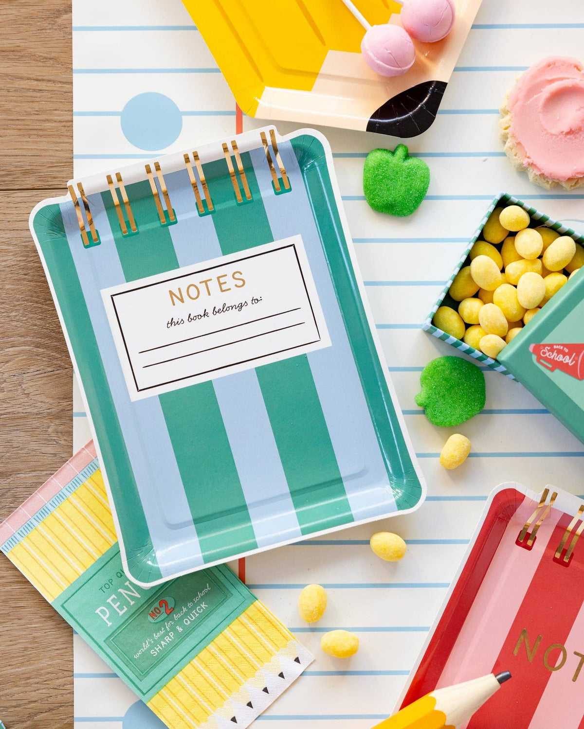 Flat lay of teal and blue striped notebook shaped paper plate on notebook paper runner with yellow candy, green apple candies, pencil shaped plate, and Back to School lunchbox treat box