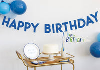 Blue puffy felt Happy Birthday banner hung above gold bar cart with birthday cake, plates, and blue balloons showing dimensional letter shadows