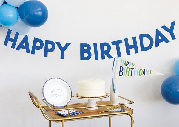 Blue puffy felt Happy Birthday banner hung above gold bar cart with birthday cake, plates, and blue balloons showing dimensional letter shadows