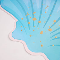 Close-up corner of the blue seashell placemat showing the pale pink scalloped edge, blue ombré ribs, and gold foil splatter.