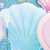 Blue seashell paper placemat under a mermaid place setting with a gold fork, iridescent mermaid-tail plate, and pink shell plate on a fish net over blue.