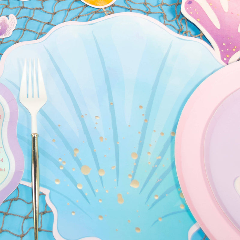 Blue seashell paper placemat under a mermaid place setting with a gold fork, iridescent mermaid-tail plate, and pink shell plate on a fish net over blue.