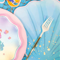 Blue seashell placemat styled with a gold fork and iridescent mermaid and shell plates on a fish-net table for an under-the-sea party.