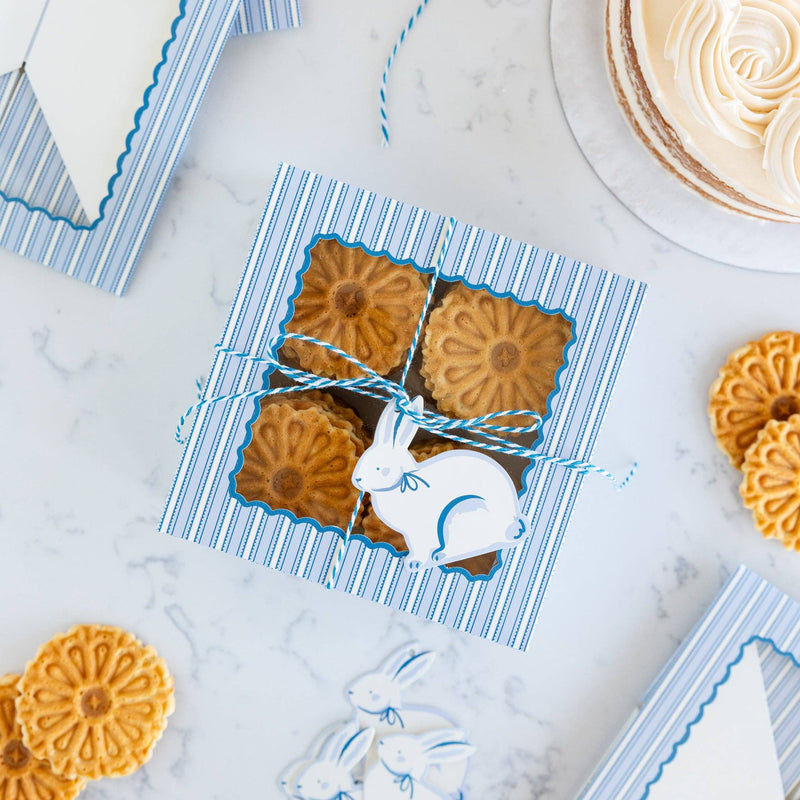 A top-down view of a blue and white striped treat box filled with flower-shaped cookies, tied with cream twine and a white bunny tag for a spring party.